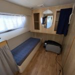Photo de la troisième et dernier cabine du bateau. On y voit à gauche un meuble de rangement situé juste devant un lit simple. Au mur, un miroir de forme ovale et deux petites étagères de rangement.