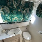 Image de la salle de bain attenante à la cabine babord. Elle est entièrement blanche avec un évier et un toilette. Un store avec des motifs tropicaux de couleur vert est installé au-dessus de l'évier
