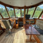 Image du salon/timonerie lumineux en bois. On voit à gauche le poste de pilotage intérieur du bateau en bois également avec un siège derrière lui et à droite une banquette avec une table en bois bordeaux.
