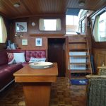 Photo de la pièce à vivre du bateau. On y voit un petit escalier du bois permettant d'accéder à la pièce. A gauche, une banquette en cuir bordeaux avec une table basse en bois et à droite un fauteuil en osier.