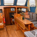Photo de la pièce à vivre du bateau tout en bois avec des murs bleus en velours. On voit à gauche le poste de pilotage intérieur en bois et à droite le côté salon avec une banquette et une table en bois dépliable.